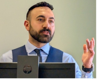 Man in a dress shirt and vest speaking behind a laptop, gesturing with one hand.