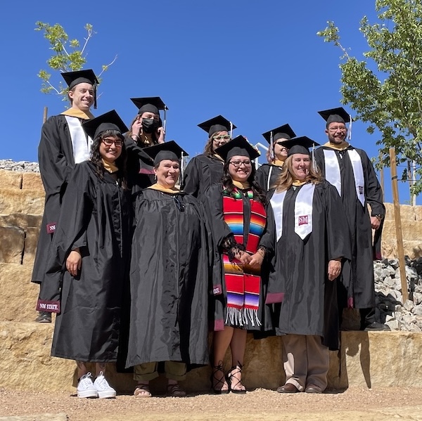 Group of graduates in black caps and gowns pose outdoors on stone steps under a clear blue sky.