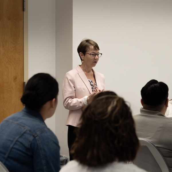 Woman in a pink blazer speaks to a seated group in a classroom or meeting room.