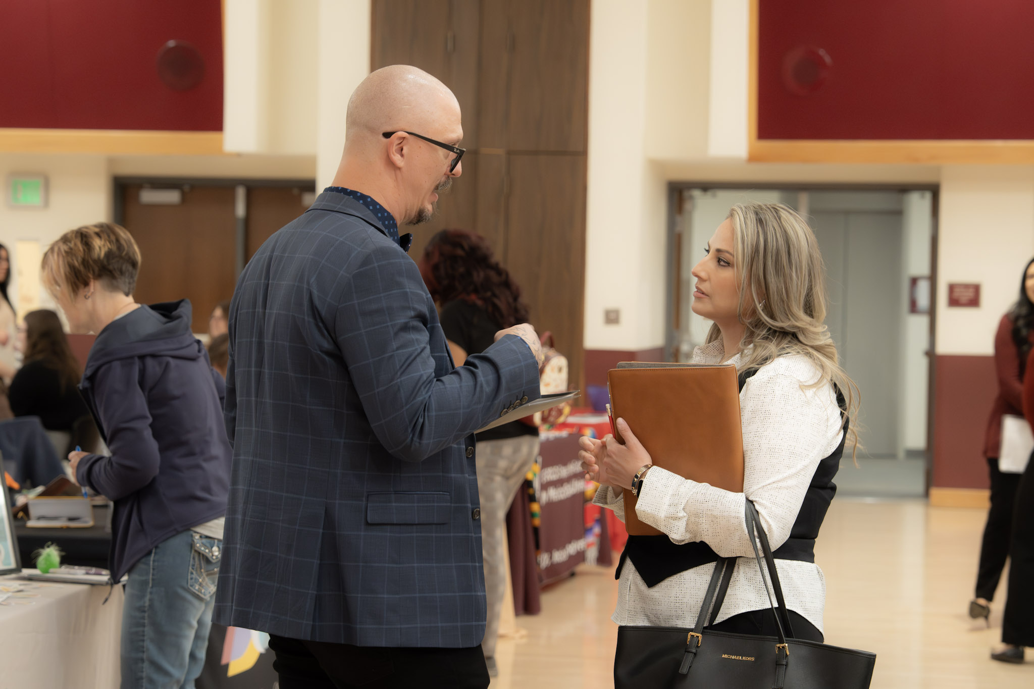 Two professionals talk at an indoor networking event near vendor tables.