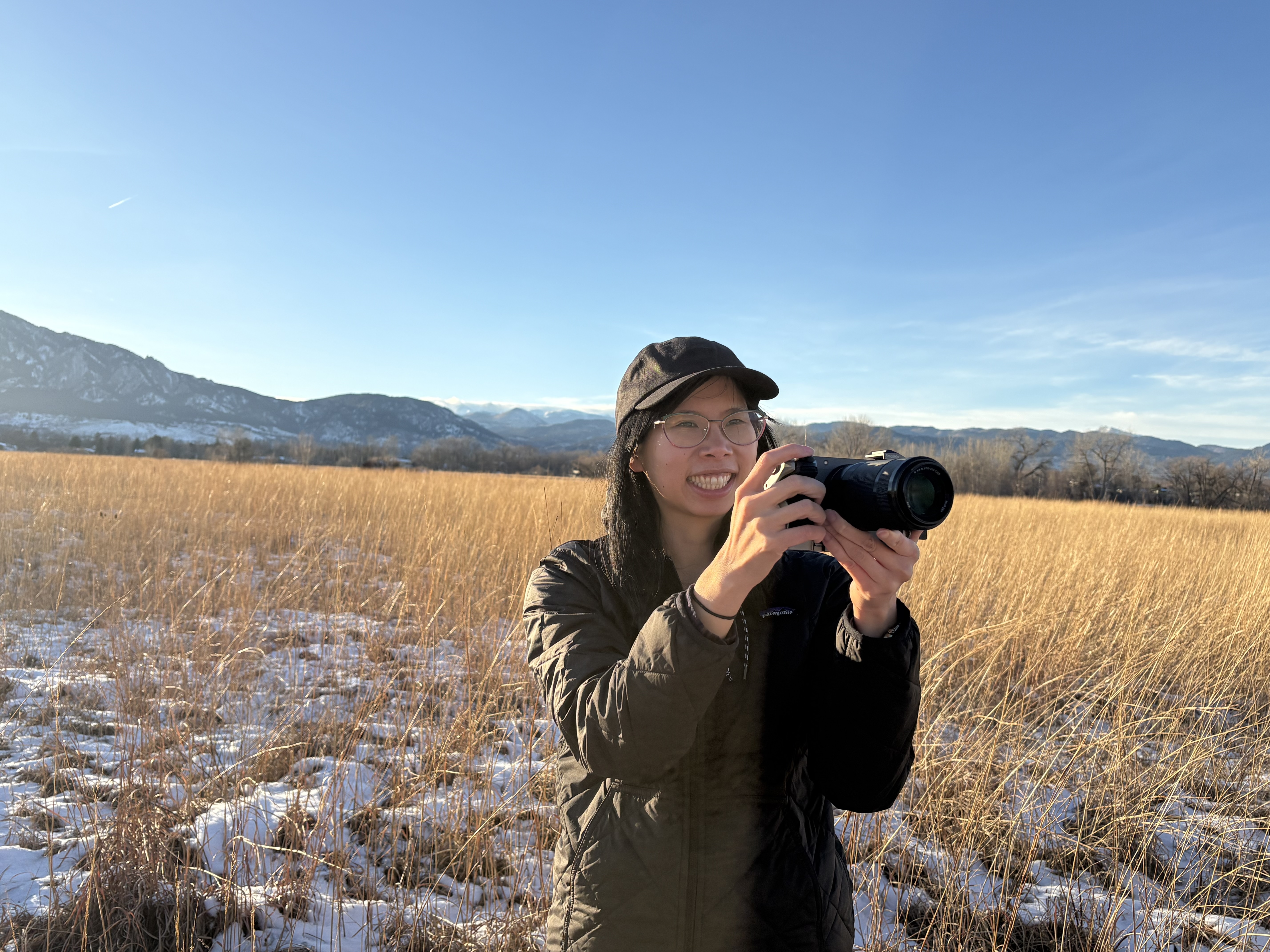 Person in a cap and glasses holds a camera in a sunlit field of tall grass with snowy patches, mountains in the distance under a clear blue sky.