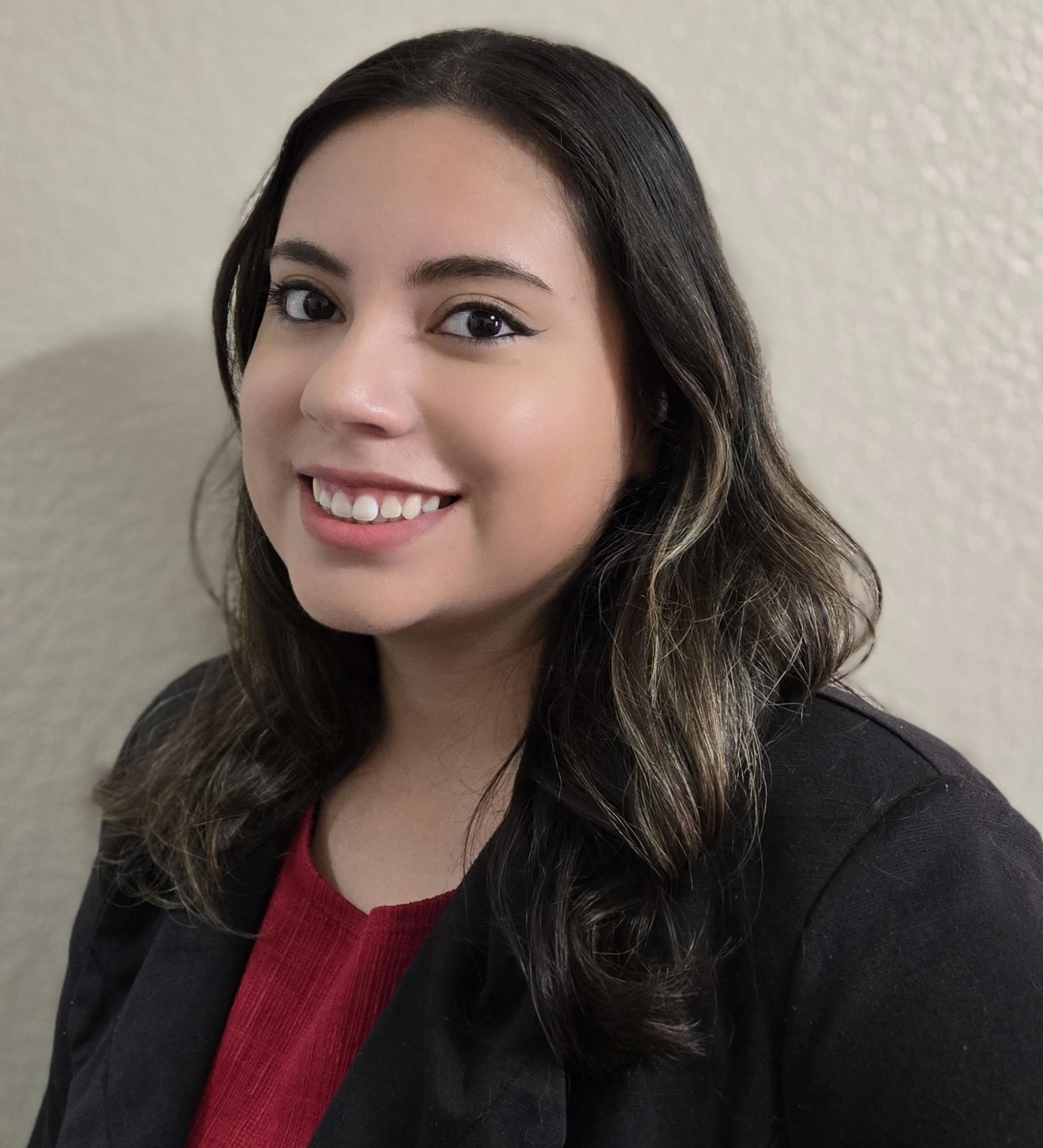 Smiling woman in a black blazer and red top against a neutral wall.