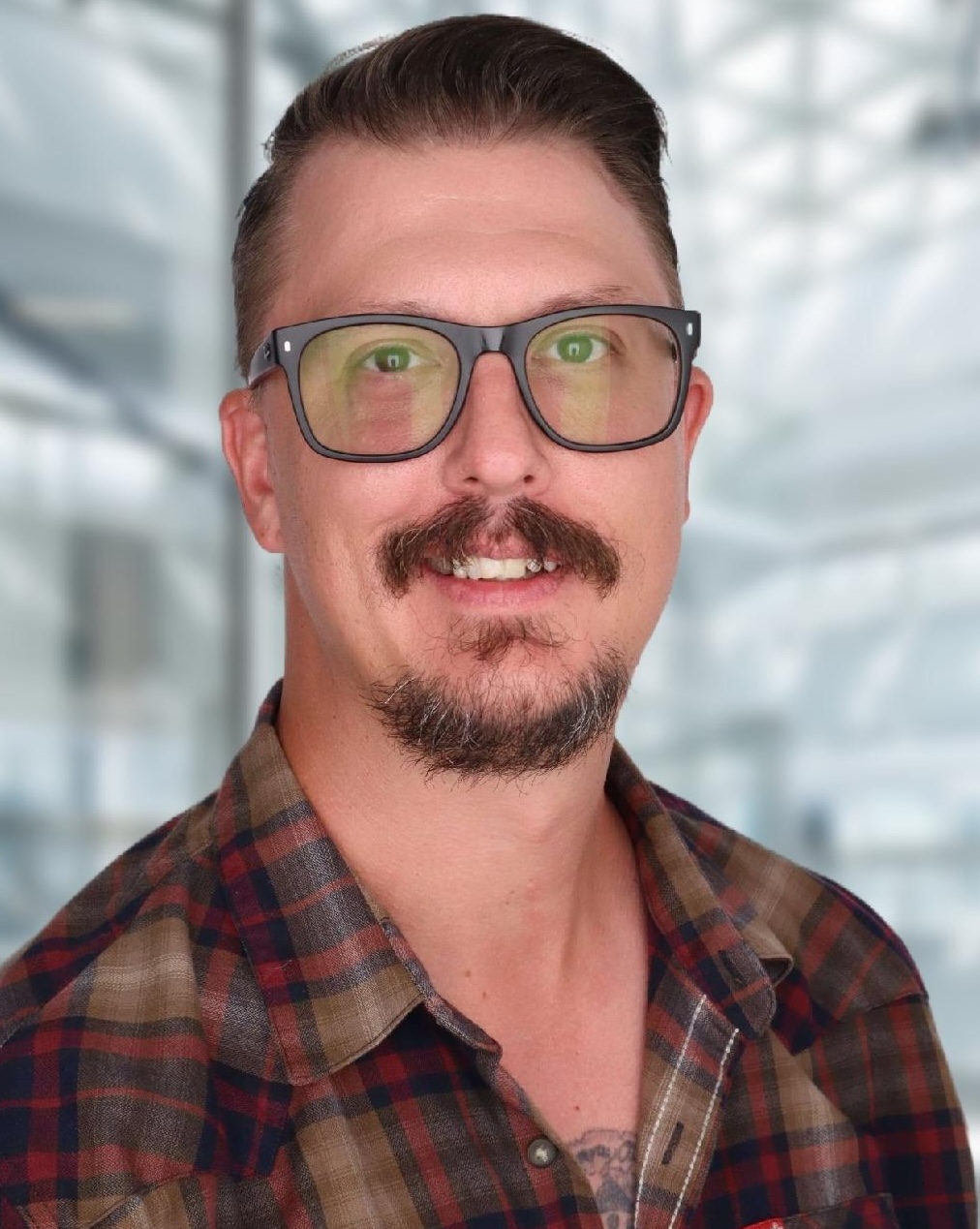 Smiling man with glasses, short hair, and a mustache wearing a plaid shirt against a blurred indoor background.