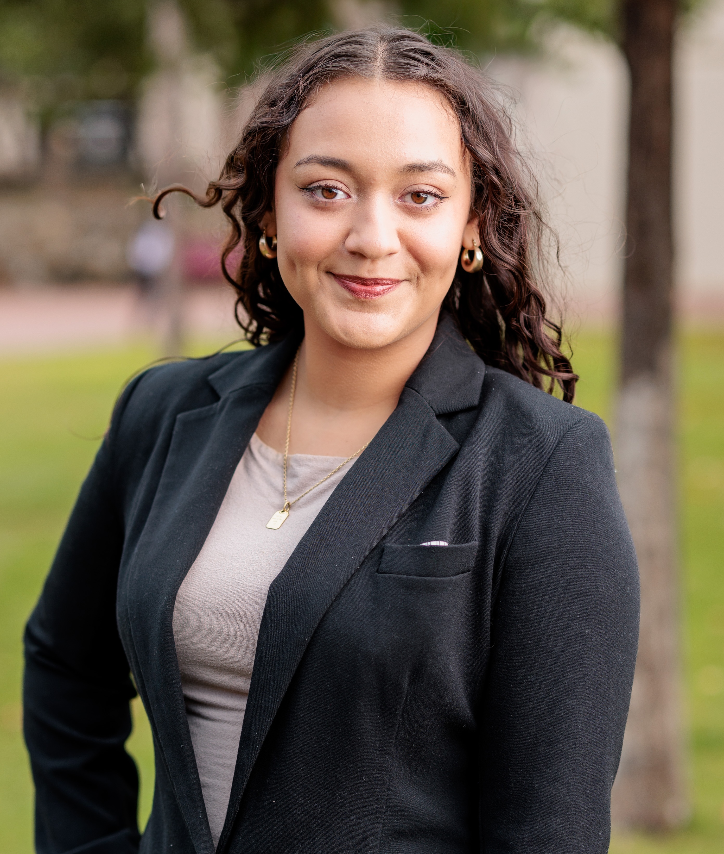 Smiling person in a black blazer and gold hoop earrings posing outdoors with a blurred background.