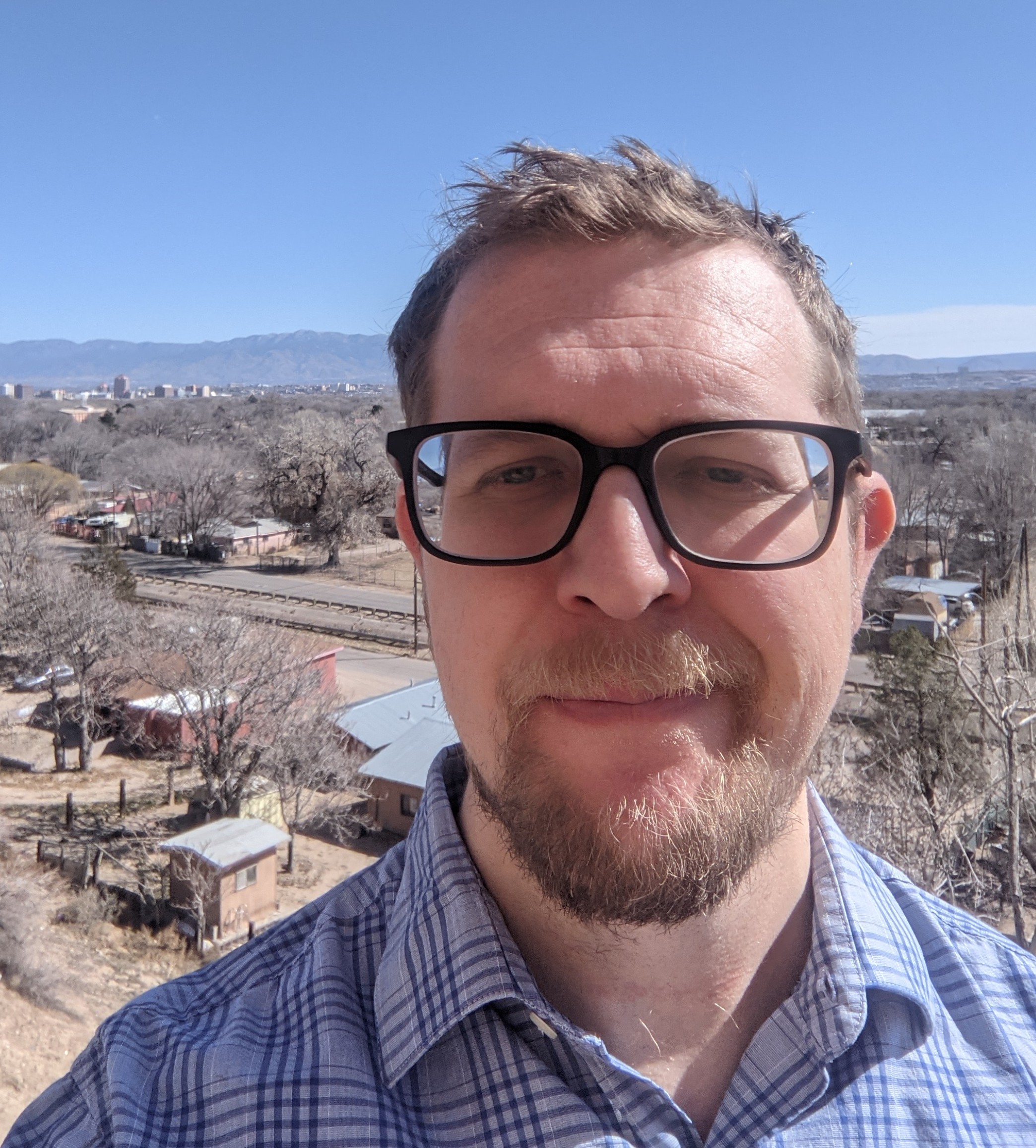 Close-up selfie of a man wearing black-framed glasses outdoors, with a small town and mountains under a clear blue sky in the background.
