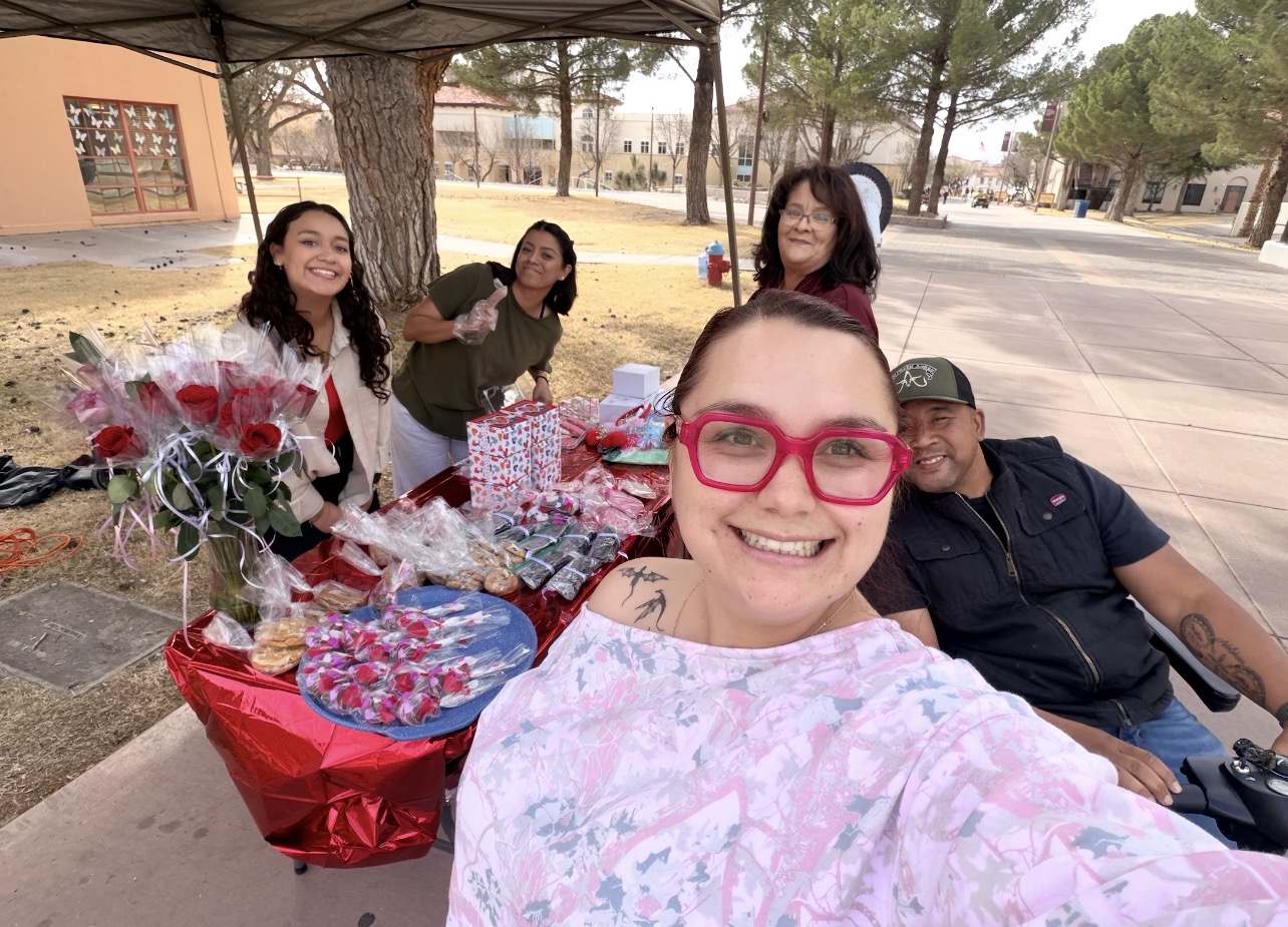 Group selfie at an outdoor table with wrapped treats and red rose bouquets under a canopy.