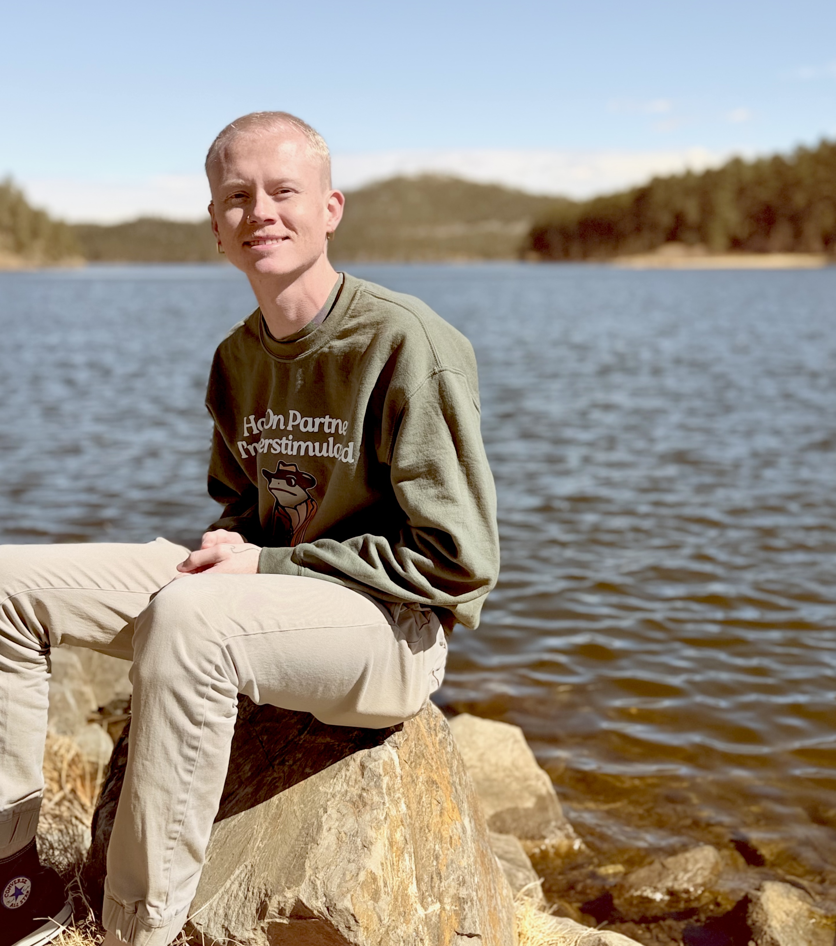 Person sitting on a large rock by a lake on a sunny day, with forested hills and blue sky in the background.