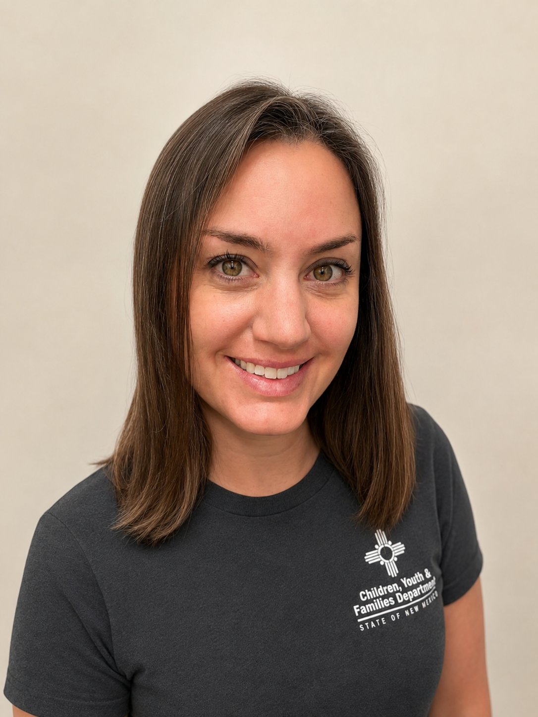 Smiling person with shoulder-length brown hair wearing a dark T-shirt with the “Children, Youth & Families Department, State of New Mexico” logo, against a plain background.
