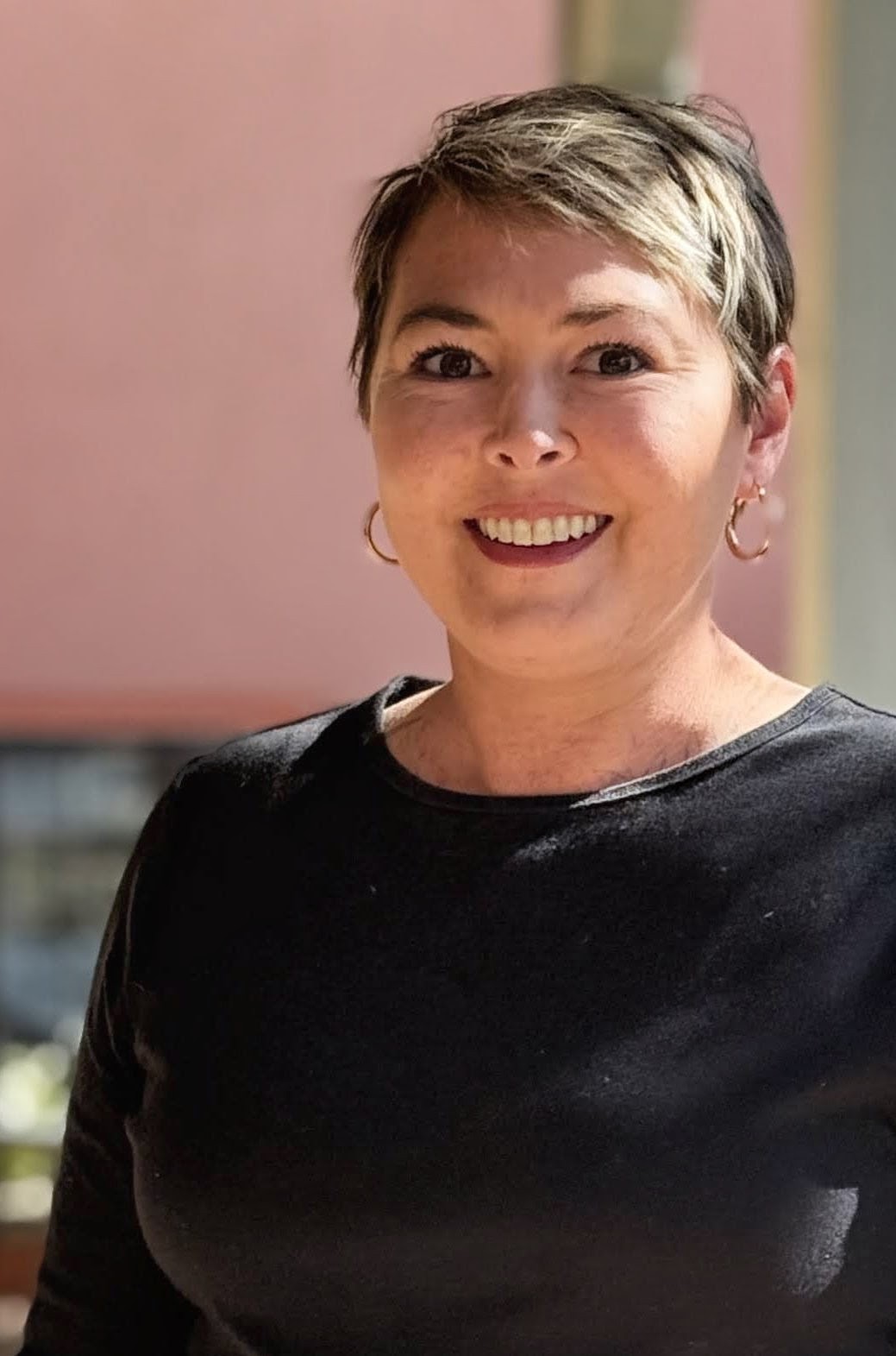 Smiling person with short highlighted hair wearing a black top and hoop earrings, standing outdoors against a softly blurred background.