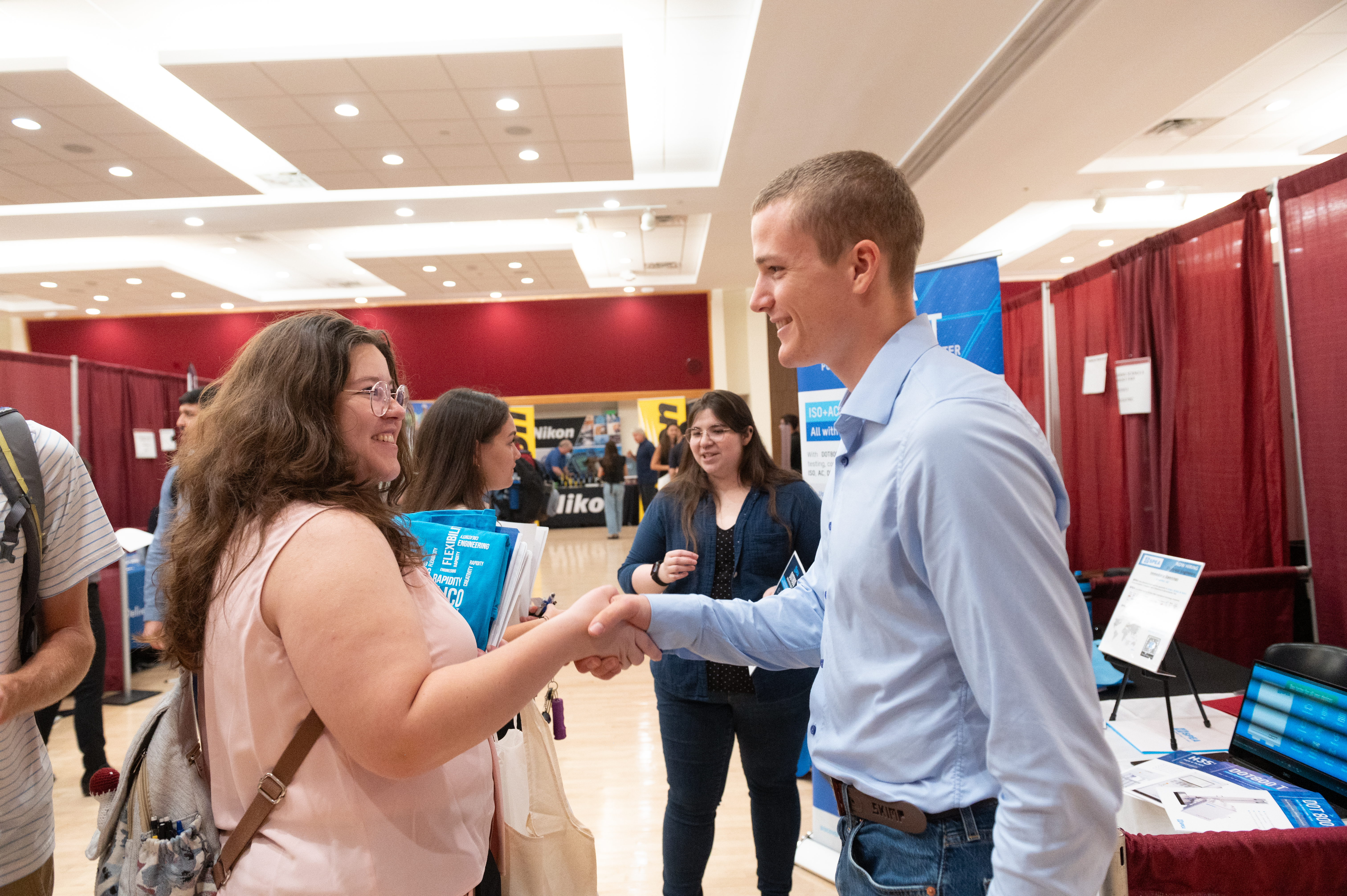 Job fair attendee shakes hands with an exhibitor at a booth in a busy conference hall.