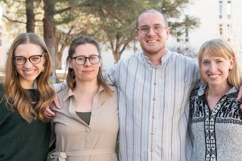 Four colleagues smiling with arms around each other outdoors on a campus.