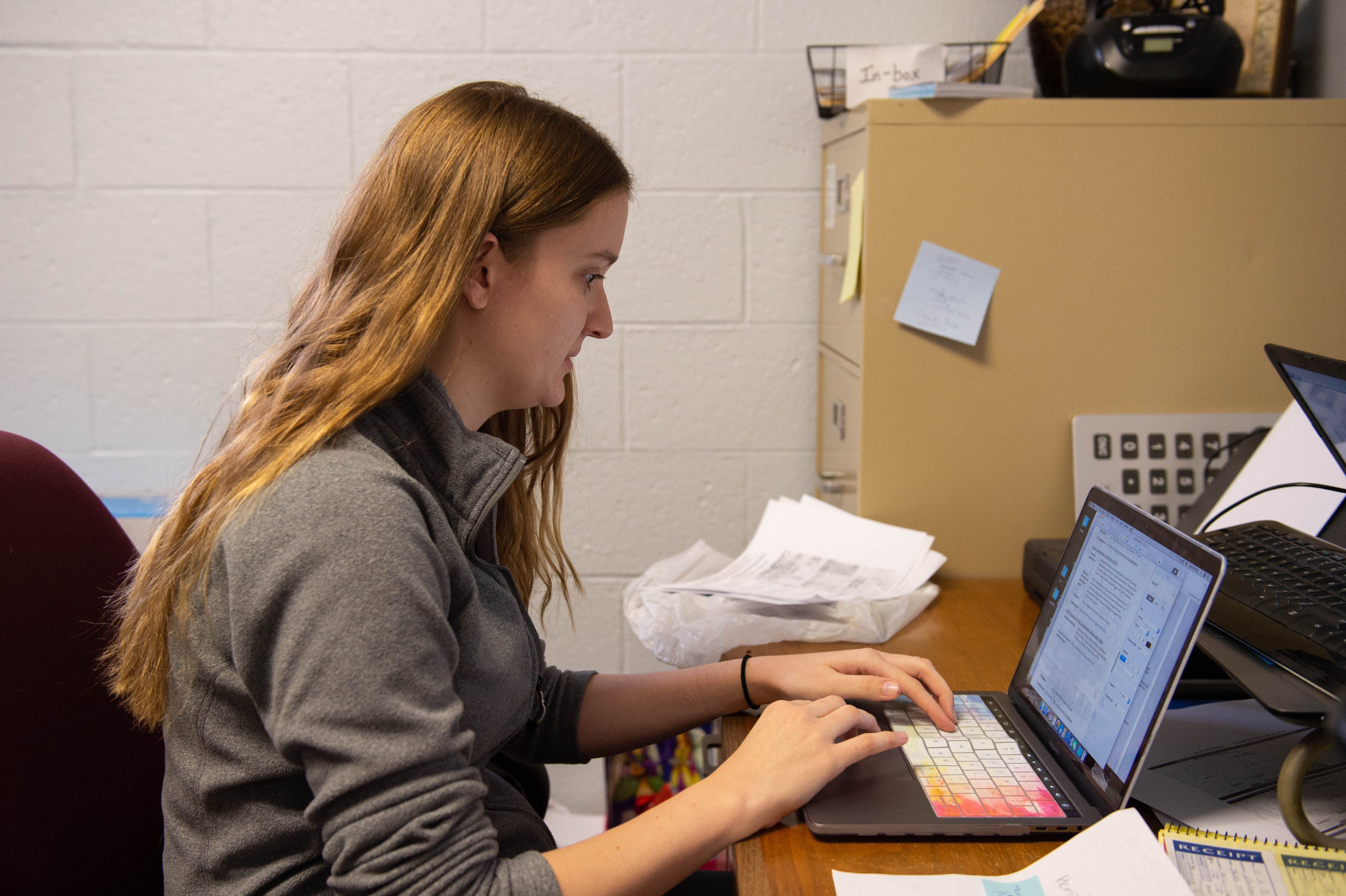 Woman working on a laptop at an office desk with papers and a filing cabinet in the background.