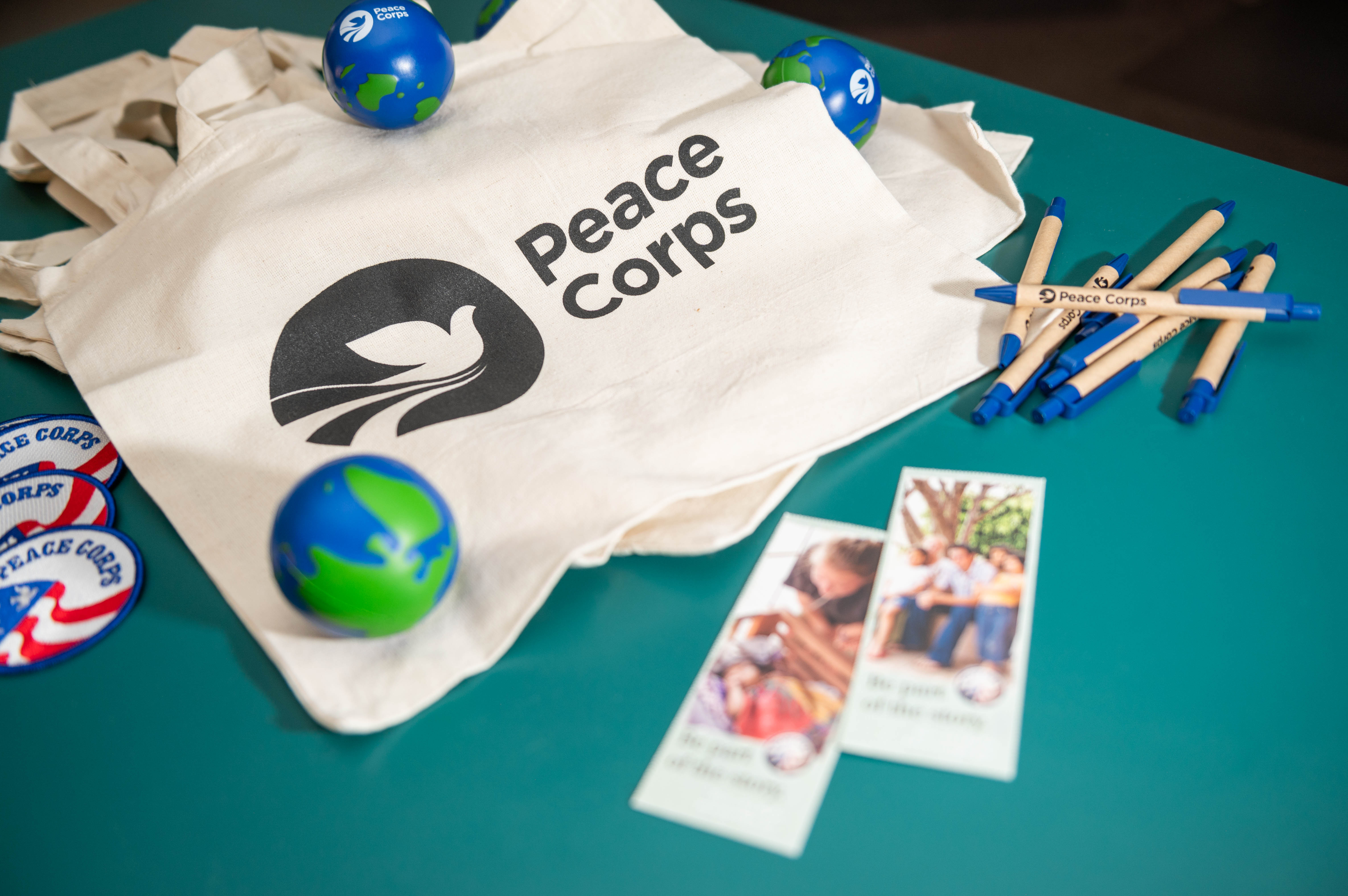 Peace Corps tote bag and promotional items on a table, including pens, patches, brochures, and globe stress balls.