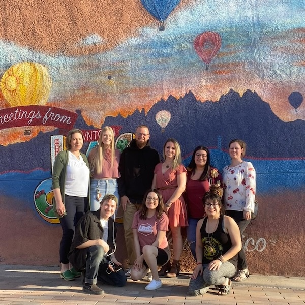 Group of people posing in front of a colorful mural with mountains, a lake, and hot air balloons.