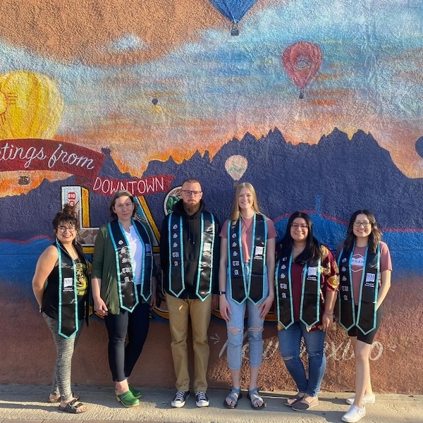 Five graduates wearing stoles stand in front of a colorful mural with mountains and hot air balloons.