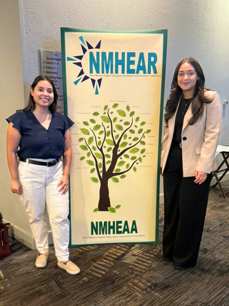 Two attendees stand on either side of a large NMHEAR conference banner featuring a tree graphic and the NMHEAA name.