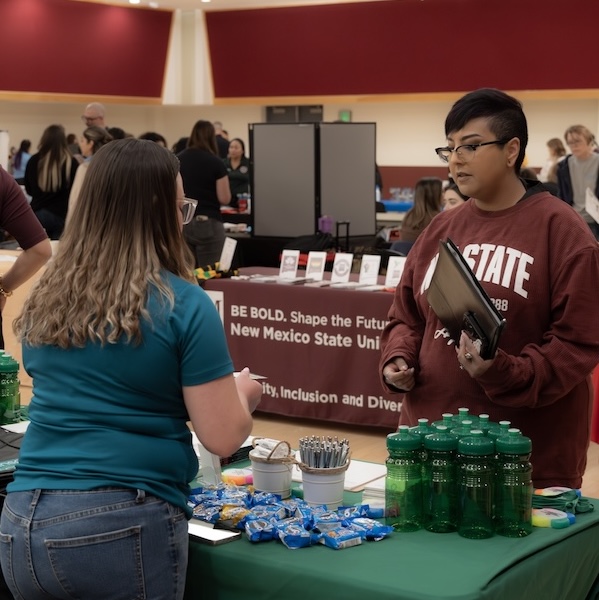 Two students talk at a campus information table with green water bottles and snacks, with a New Mexico State University banner in the background.