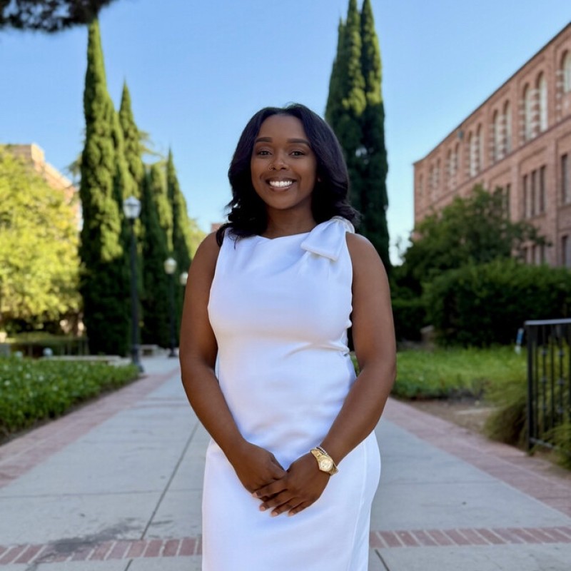 Woman in a sleeveless white dress standing on a campus walkway lined with tall trees, smiling at the camera.