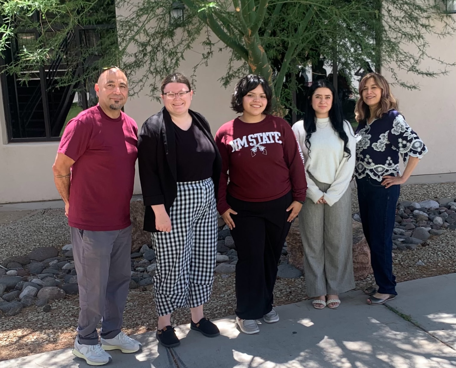 Five people standing outdoors together in front of a building and trees, posing for a group photo.
